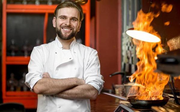 Smiling chef with arms crossed standing in front of a flaming skillet